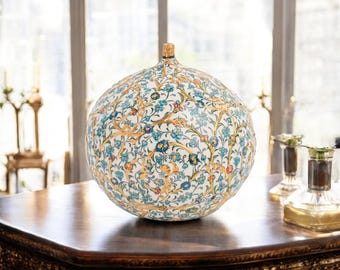 Può includere: Un vaso sferico in ceramica con un elaborato motivo floreale in blu, arancione e oro su sfondo bianco. Il vaso è appoggiato su un tavolo di legno scuro. Il vaso ha una piccola sommità dorata.