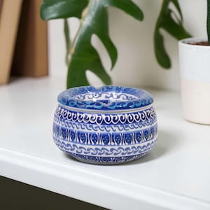 Puede incluir: Un cenicero redondo de cerámica con un patrón floral azul y blanco. El cenicero tiene un borde ancho y un diseño detallado alrededor del cuerpo. La paleta de colores incluye tonos de azul y blanco.