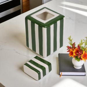 Pu&ograve; includere: Scatola di fazzoletti quadrata in ceramica bianca con strisce verdi verticali. Il coperchio &egrave; una versione pi&ugrave; piccola della scatola, anch'esso con strisce verdi. Un piccolo vaso bianco con fiori e un taccuino nero sono accanto.