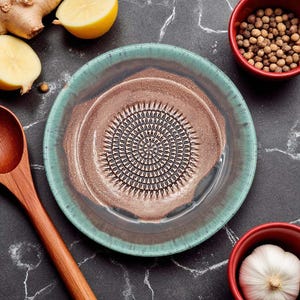 Puede incluir: Un plato rallador de cerámica con borde turquesa y centro marrón, con superficie de rallado texturizada. El plato está rodeado de ingredientes como jengibre, limón, granos de pimienta y ajo, lo que sugiere uso culinario.