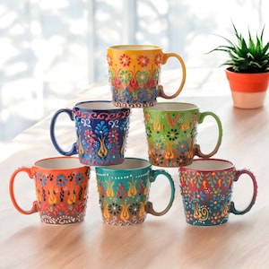 Pode incluir: Seis canecas de cerâmica empilhadas com padrões florais coloridos. As canecas apresentam tons de laranja, azul, verde e amarelo, com detalhes brancos e castanhos. Cada caneca tem uma alça curva.