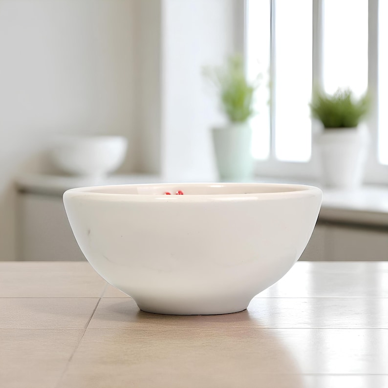 May include: A white ceramic bowl with a rounded shape. The bowl is sitting on a light-colored surface. The background includes a window and a glimpse of a kitchen counter. Small red items are visible inside the bowl.