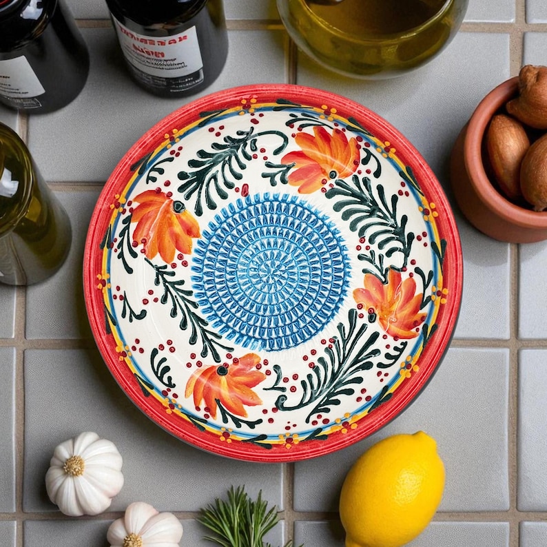 May include: A decorative ceramic plate with a red rim, featuring a floral design with orange flowers, blue center, and green leaves. The plate is surrounded by ingredients like garlic, lemons, and rosemary.