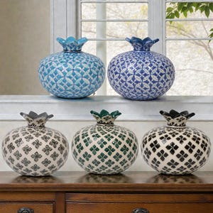 Può includere: Sei vasi decorativi in ceramica a forma di melograno. Due sono blu chiaro e scuro, e quattro sono bianchi con motivi floreali neri, verdi e grigi. I vasi hanno una superficie strutturata e sono esposti su una superficie di legno.