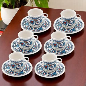 May include: A six-piece coffee set with white ceramic cups and saucers. Each piece is decorated with a blue, red, and white floral design. The cups have small handles and sit on matching saucers. The set is displayed on a dark brown surface.