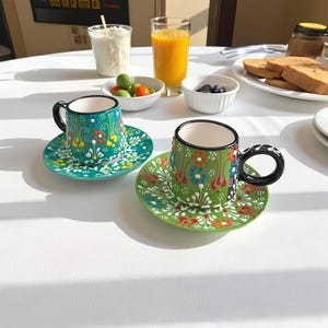 May include: Two ceramic coffee cups and saucers with colorful floral designs. The cups are green with black handles and rims. The saucers are green with white floral patterns. The cups and saucers are on a white surface.