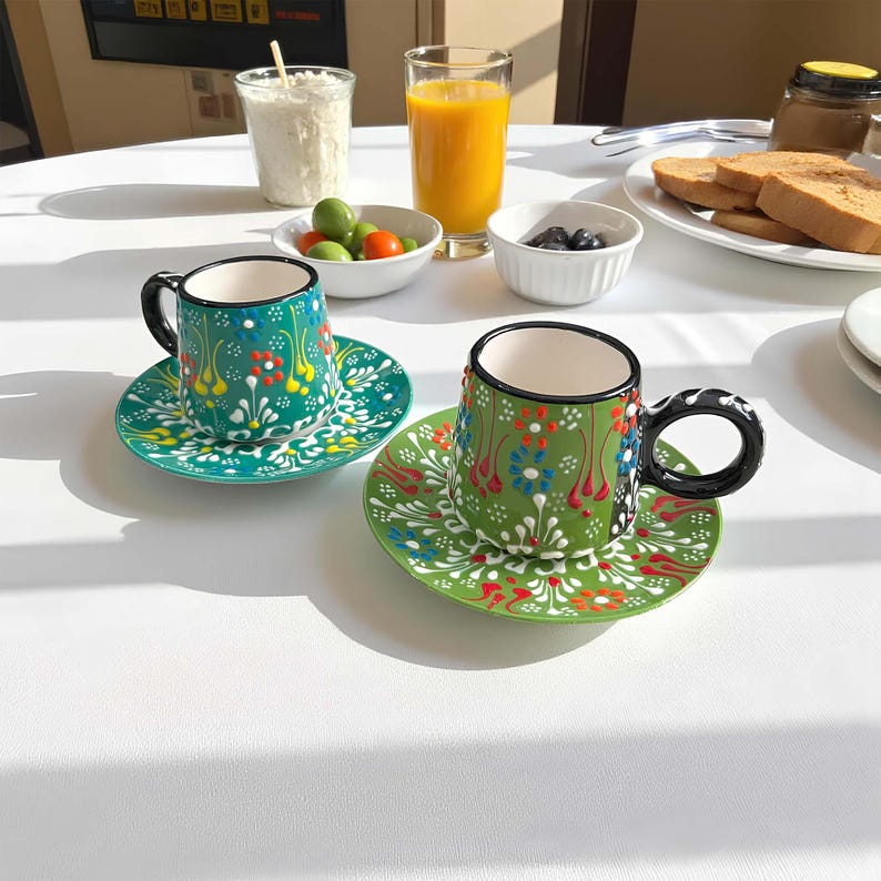May include: Two ceramic coffee cups and saucers with colorful floral designs. The cups are green with black handles and rims. The saucers are green with white floral patterns. The cups and saucers are on a white surface.