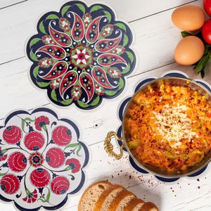 Könnte beinhalten: Drei dekorative Untersetzer mit gewelltem Rand und aufwendigen Blumen- und Paisley-Designs in Rot, Grün und Weiß auf marineblauem Hintergrund. Ein Untersetzer hält eine Metallpfanne mit einem gekochten Gericht. Geschnittenes Brot und Eier sind ebenfalls sichtbar.