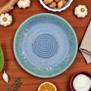 Puede incluir: Un plato de cerámica azul y turquesa con una superficie texturizada y un diseño en espiral en el centro. El plato está rodeado de ingredientes como ajo, nueces y una rodaja de naranja sobre una superficie de madera.