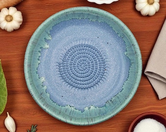 Puede incluir: Un plato de cerámica azul y turquesa con una superficie texturizada y un diseño en espiral en el centro. El plato está rodeado de ingredientes como ajo, nueces y una rodaja de naranja sobre una superficie de madera.
