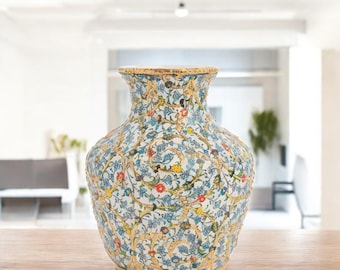 Può includere: Un vaso decorativo in ceramica con un motivo floreale dettagliato in blu, rosso, giallo e verde su sfondo bianco. Il vaso ha un corpo largo e un collo più stretto, con un bordo e una base dorati. Il vaso è appoggiato su un tavolo di legno.