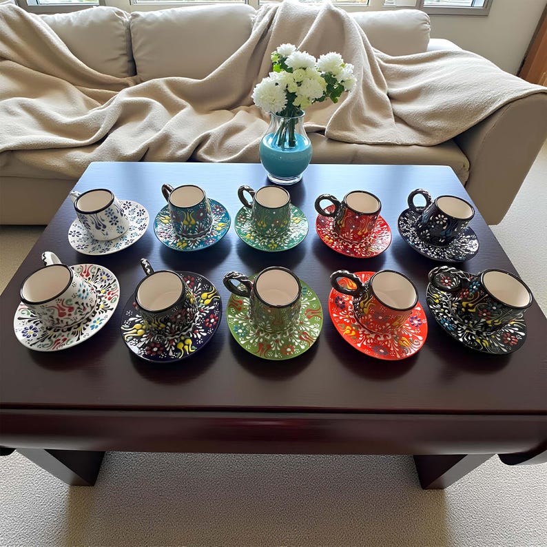 May include: A collection of colorful, hand-painted ceramic coffee cups and saucers with intricate floral designs. The cups are in various colors, including green, red, and black, and are arranged on a dark brown table. A vase of white flowers sits nearby.