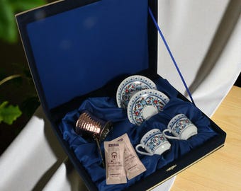 May include: A blue velvet-lined gift box containing two small coffee cups and saucers with a floral design, a copper coffee pot, and two packets of coffee. The box is closed with a gold clasp.