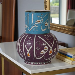 Puede incluir: Un jarrón decorativo de cerámica de dos piezas con caligrafía árabe. La sección inferior es de color púrpura oscuro con escritura blanca y detalles en naranja. La sección superior es azul claro con escritura blanca. El jarrón está sobre una mesa blanca.