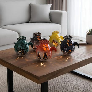 May include: A collection of five ceramic teapot-shaped candle holders in various colors, including green, black, orange, yellow, and dark brown. Each has a decorative top and cut-out floral designs, casting light onto the wooden coffee table.