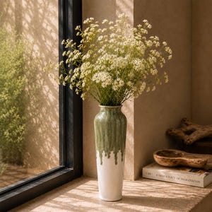 Könnte beinhalten: Eine hohe Keramikvase mit konischer Silhouette, die eine glänzende salbeigrüne Glasur aufweist, welche über eine einfarbig weiße Basis läuft. Die Vase ist mit einem großen, luftigen Strauß kleiner weißer Schleierkrautblüten mit dünnen grünen Stielen gefüllt.