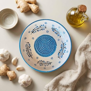 Puede incluir: Un plato rallador de cerámica blanca con centro y borde azules, decorado con diseños de hojas negras y azules. El plato está rodeado de dientes de ajo, jengibre, un cuenco pequeño y una botella de aceite de oliva.
