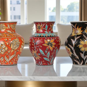 May include: Three decorative ceramic vases with floral patterns. The vases are orange, red, and black, each with a unique design featuring flowers and leaves. They sit on a white marble surface, showcasing their intricate details.
