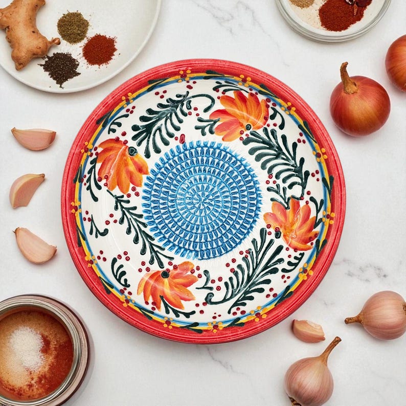 May include: A decorative ceramic plate with a red rim, featuring a floral design in orange, blue, and green. The plate is surrounded by spices, garlic cloves, and onions, suggesting a culinary theme.