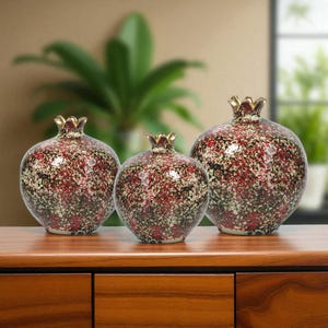 May include: Three ceramic pomegranate sculptures with a speckled red, black, and cream pattern. Each has a gold-coloured crown. The pomegranates are arranged on a wooden surface, with a blurred green plant in the background.