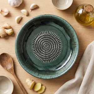 Puede incluir: Un cuenco rallador de ajo de cerámica verde azulado con un centro texturizado. El cuenco está rodeado de dientes de ajo, jengibre, una cuchara de madera, cuencos pequeños, aceite de oliva y un paño de lino, todo sobre una superficie de madera.