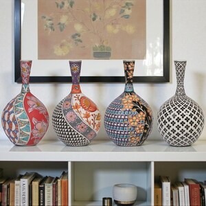 May include: Four decorative ceramic vases with patterned designs in red, black, blue, and white. The vases are displayed on a white shelf with books and a framed artwork in the background. The vases have a bulbous shape.