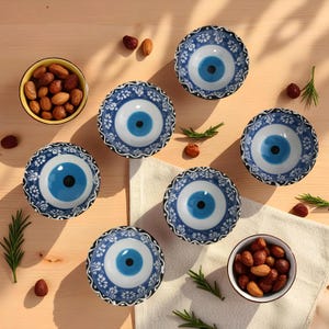 Puede incluir: Vista cenital de seis cuencos pequeños y redondos de cerámica blanca y azul con un diseño de ojo. Los cuencos están dispuestos sobre una superficie de madera clara con dos cuencos pequeños de frutos secos, ramitas de romero y frutos secos dispersos.