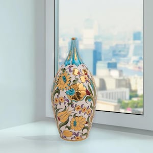 Op de afbeelding: Een decoratieve vaas met een crème achtergrond en een bloemenpatroon in goud, blauw en geel. De vaas heeft een slanke hals en een brede body. De bovenkant van de vaas is turquoise. De vaas staat bij een raam.