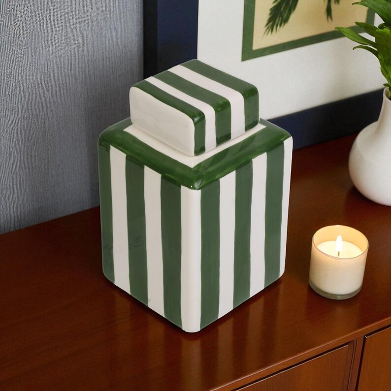 Pu&ograve; includere: Un vaso decorativo in ceramica con coperchio, a righe verticali verdi e bianche. Il vaso &egrave; rettangolare con un coperchio quadrato. Una candela accesa in un portacandele di vetro &egrave; nelle vicinanze.