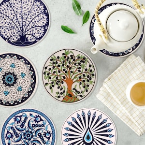 Puede incluir: Un conjunto de posavasos de cerámica con diseños intrincados. Los posavasos presentan patrones en azul y blanco, incluyendo motivos florales, de árboles y de plumas de pavo real. Una tetera blanca y una taza de té también están presentes, sugiriendo un ambiente temático de té.