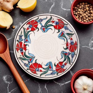 Puede incluir: Un plato de cerámica blanco con un diseño floral con detalles en rojo, azul y verde. El plato tiene un borde negro y está rodeado de ingredientes como jengibre, limón y especias.