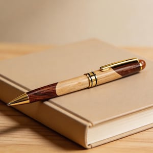 May include: A handcrafted wooden pen with a gold-colored nib and accents rests on a closed beige book. The pen features a combination of light and dark wood, with a gold clip and a rounded end. The pen is a writing instrument.