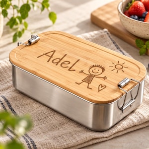 Könnte beinhalten: Eine rechteckige Brotdose aus Edelstahl mit einem Bambusdeckel. Auf dem Deckel ist der Name "Adel" eingraviert, zusammen mit einer einfachen Strichzeichnung einer Person, einer Sonne und einem Herz. Zwei Metallverschlüsse sichern den Deckel.