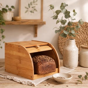 May include: A rectangular bamboo bread box with a roll-top lid and a small wooden knob. Inside the open bread box sits a loaf of dark, crusty bread topped with sunflower seeds. The bread box is constructed from light-colored, natural bamboo.