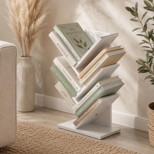 May include: A white, tree-shaped bookshelf with multiple angled shelves holding various books. The structure features a central vertical spine with branches extending outward to support books titled Kinfolk, This is Home, and Nordic Interiors Homes.