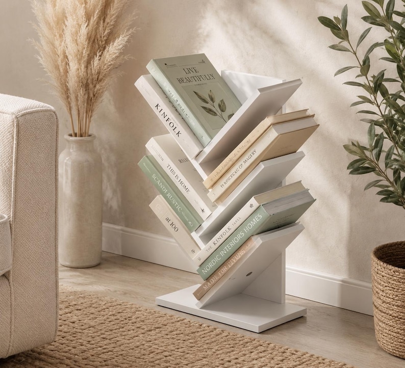 May include: A white, tree-shaped bookshelf with multiple angled shelves holding various books. The structure features a central vertical spine with branches extending outward to support books titled Kinfolk, This is Home, and Nordic Interiors Homes.
