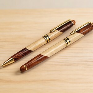 May include: Two wooden pens with gold-colored accents. The pens have a light wood and dark wood design. The pen tips and clips are gold. The pens are lying on a light wood surface.
