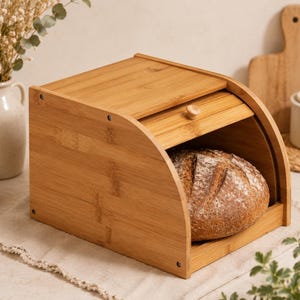 Könnte beinhalten: Ein hellbrauner Brotkasten aus Bambus mit Rolldeckel und einem kleinen hölzernen Knaufgriff. Der Kasten hat ein geschwungenes Seitenprofil und enthält einen runden, krustigen Laib Brot mit einer eingeritzten Oberfläche im Inneren.