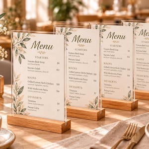 Könnte beinhalten: Vier vertikale Menükarten in durchsichtigen Acrylhaltern mit hellen Holzsockeln. Jede Karte hat einen weißen Hintergrund mit Aquarell-Blattillustrationen, dem Wort Menu sowie Abschnitten für Vorspeisen, Hauptgerichte und Desserts.