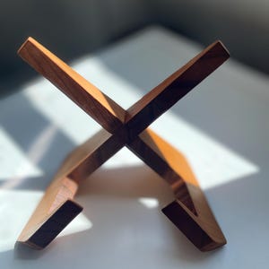 Könnte beinhalten: Ein hölzerner Buchständer mit einem X-förmigen Design. Der Ständer ist aus hellbraunem Holz gefertigt und hat eine schlichte, moderne Ästhetik. Der Ständer ist für Bücher oder andere Gegenstände konzipiert.