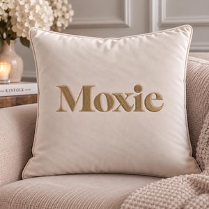 May include: A square cream-colored throw pillow featuring the word Moxie embroidered in a metallic gold thread across the center. The pillow has a smooth, soft-touch fabric finish and is finished with matching cream-colored piping along the edges.