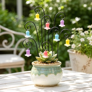 Può includere: Un vaso in ceramica con smalto a goccia verde e bianco contiene una pianta succulenta. Steli in filo metallico nero con estremità ricurve si estendono dal centro, ognuno dei quali sostiene un piccolo fiore traslucido a forma di campana giallo, rosa, blu o viola.