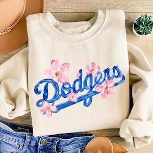 May include: A cream-colored crewneck sweatshirt featuring the word Dodgers in a blue script font with a watercolor-style underline. The design is decorated with several pink cherry blossom flowers scattered around and overlapping the blue text.