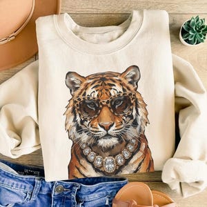 May include: A cream-colored crewneck sweatshirt featuring a graphic illustration of a tiger wearing leopard-print sunglasses and a large, chunky rhinestone necklace. The tiger has orange and black fur with white accents around the muzzle.