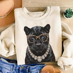 May include: A cream-colored crewneck sweatshirt featuring a graphic illustration of a black panther wearing leopard-print glasses and a large, multi-faceted crystal statement necklace. The panther has detailed whiskers and piercing eyes.