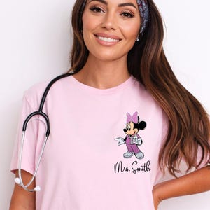 以下が含まれることがあります： 看護師の衣装を着たミニーマウスのグラフィックが特徴のピンクのTシャツ。筆記体で「Mrs. Smith」と書かれています。Tシャツはクルーネックと半袖です。聴診器が首にかけられています。