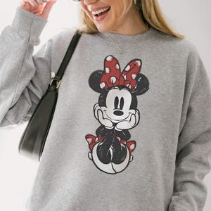 May include: A gray sweatshirt featuring a distressed graphic of Minnie Mouse with a red bow and accents. The sweatshirt is long-sleeved and has a crew neck. The overall style is casual and comfortable.