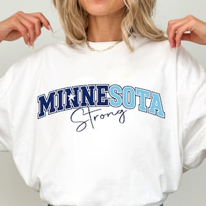 May include: White t-shirt with the text "MINNESOTA Strong" in blue. The word "MINNESOTA" is in a block letter style, and "Strong" is in a script font. The shirt is made of a soft material.