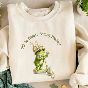 Könnte beinhalten: Cremefarbenes Sweatshirt mit einer skurrilen Grafik einer Froschfigur auf einem Roller, die einen Hut mit Hasenohren trägt und Blumen hält. Der Text "Off to Commit Spring Foolery" ist über dem Froschbogen angeordnet. Das Sweatshirt hat einen Rundhalsausschnitt und lange Ärmel.