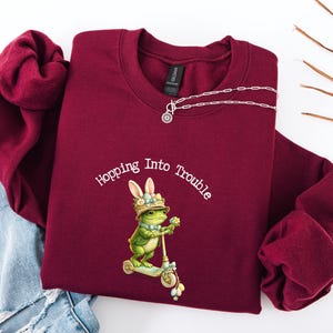 Peut inclure: Sweat-shirt bordeaux à col rond avec l'inscription "Hopping Into Trouble" et un dessin d'une grenouille avec des oreilles de lapin sur un scooter. Un collier argenté est posé sur le sweat-shirt.